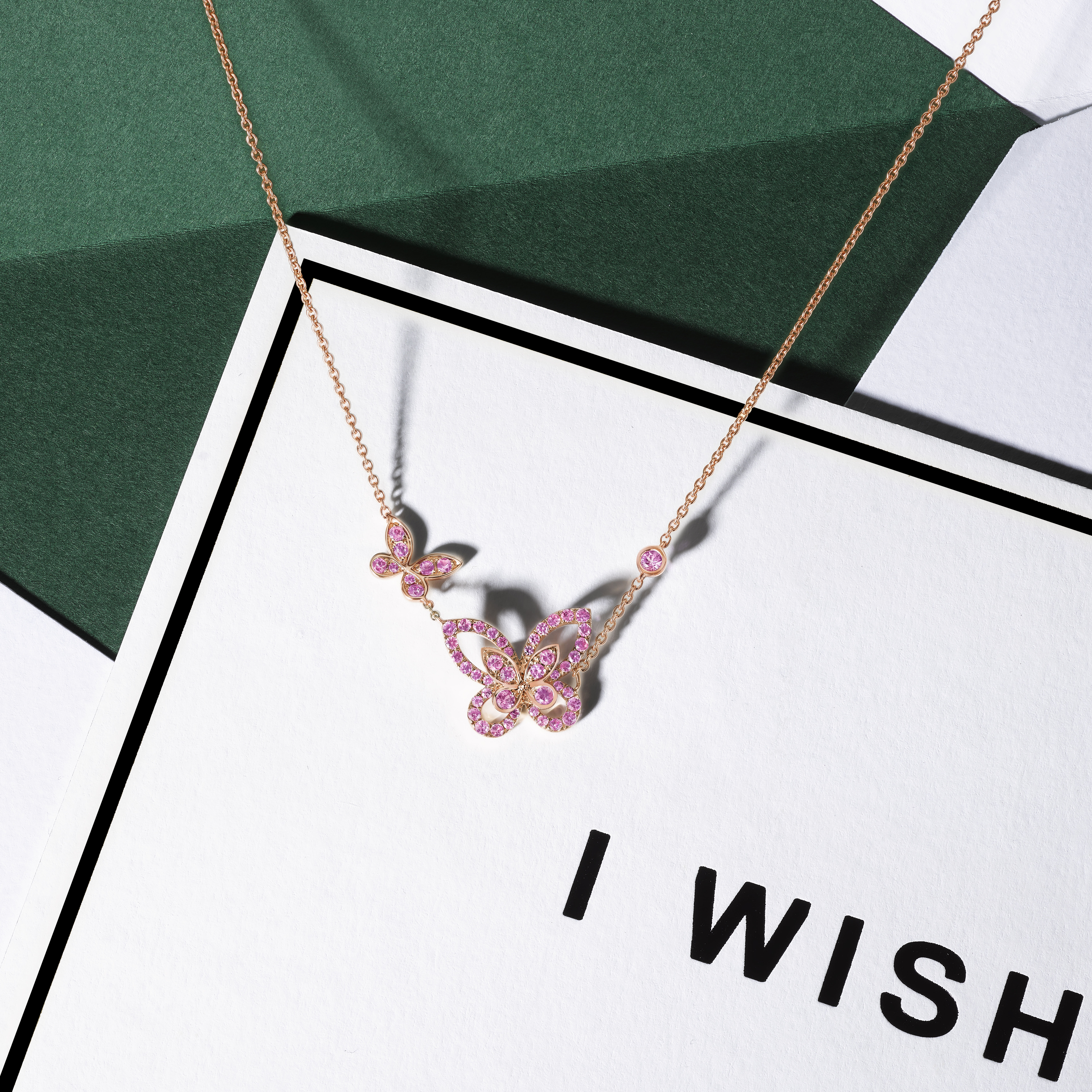 Still life image of Graff diamond collection Rose Gold and Pink Sapphire Butterfly Silhouette necklace positioned on an 'I WISH' festive greetings card 