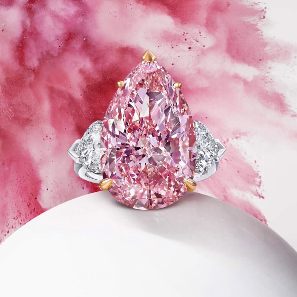A Graff ring featuring a Fancy Vivid Pink Internally Flawless pear shape diamond and heart shape white diamond shoulder stones with pink explosion background