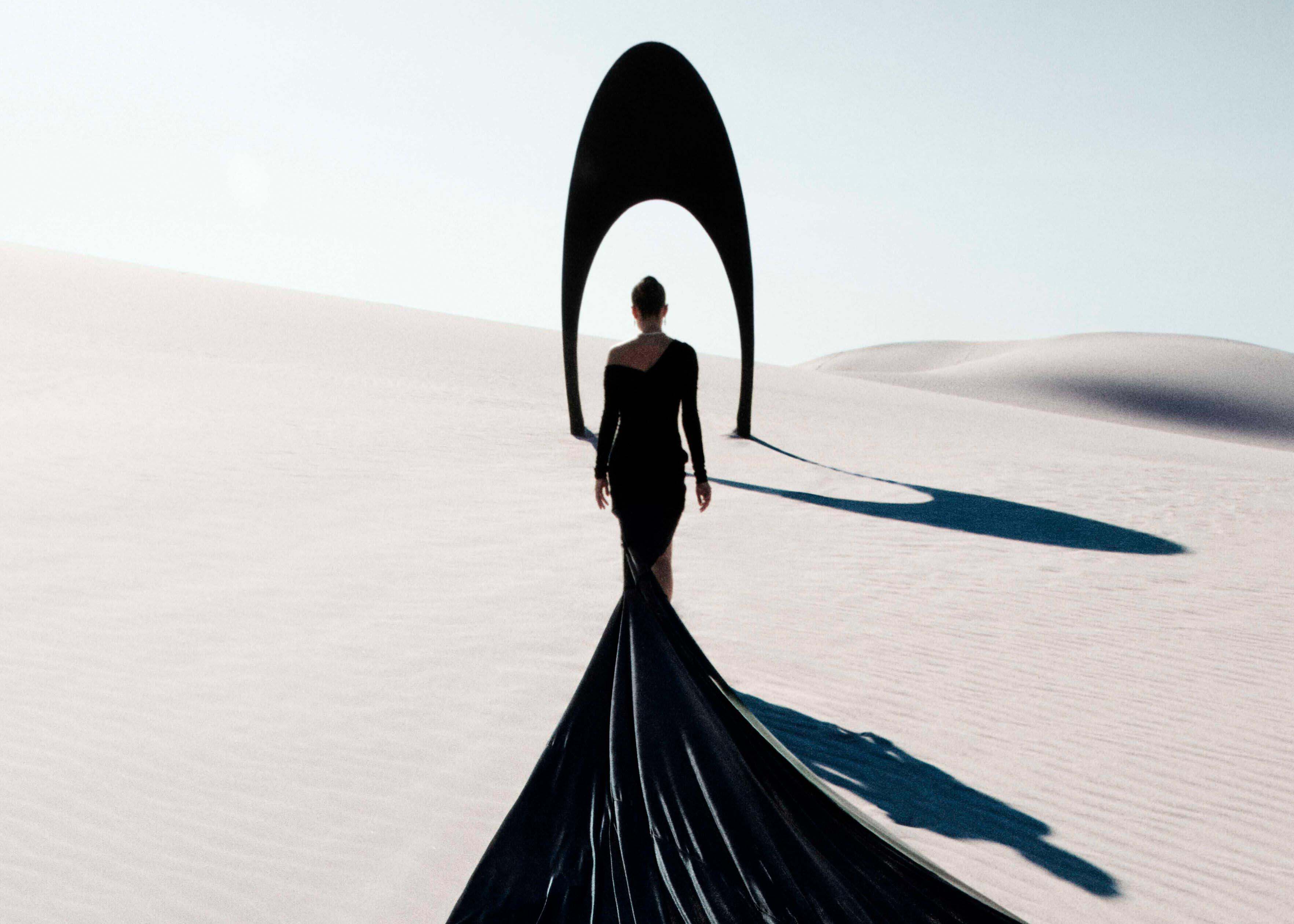 Close up of a model wearing Graff Night Moon emerald and diamond high jewellery earrings and necklace from the Tribal jewellery collection, in a desert in front of a black sculpture